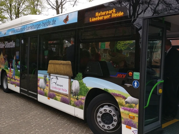 HeideShuttle Bus mit Landschaftsmotiven, Schafen und Heide beklebt. Tür offen, Fahrgäste steigen ein.