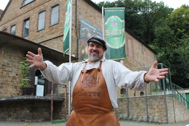 Barres Brauwelt Eine fröhliche Person in traditioneller Bierbrauerkleidung steht mit offenen Armen vor Barres Brauwelt.