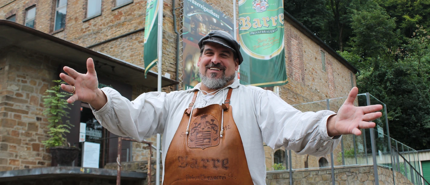 Barres Brauwelt Eine fröhliche Person in traditioneller Bierbrauerkleidung steht mit offenen Armen vor Barres Brauwelt.