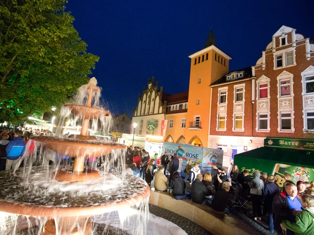 Lübbecke_Bierbrunnenfest2_Oliver Krato.jpg Festliches Stadtleben bei Nacht mit beleuchtetem Bierbrunnen, umringt von feiernden Menschen.