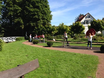 Kurpark Bad Holzhausen Grüner Park mit Outdoor-Fitnessgeräten, Menschen beim Training und ein weißes Gebäude im Hintergrund.