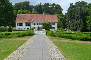 Zweigeschossiges Gebäude mit rotem Satteldach, umgeben von Bäumen und Rasenflächen im Vordergrund.