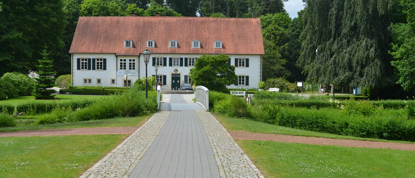 Zweigeschossiges Gebäude mit rotem Satteldach, umgeben von Bäumen und Rasenflächen im Vordergrund.