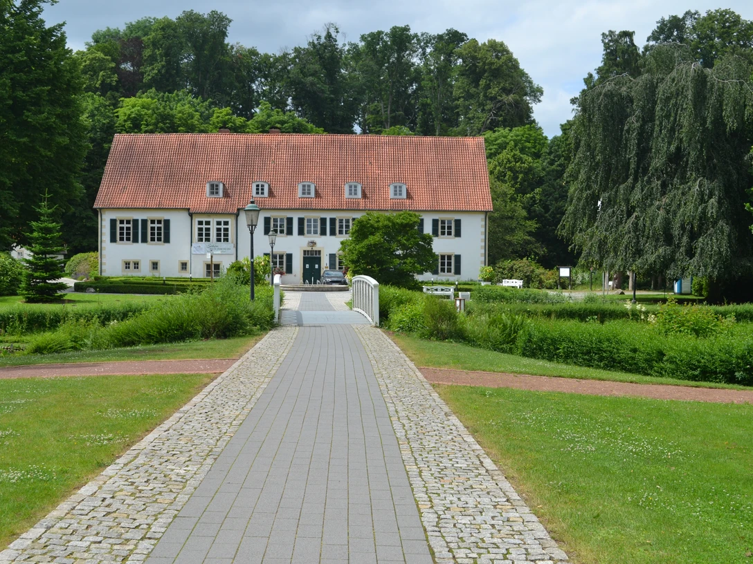 Pr-Oldendorf - Haus des Gastes Zweigeschossiges Gebäude mit rotem Satteldach, umgeben von Bäumen und Rasenflächen im Vordergrund.
