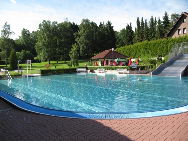 Freibad mit breiten Treppen, großer Wasserrutsche und umgeben von Bäumen und grünen Wiesen.