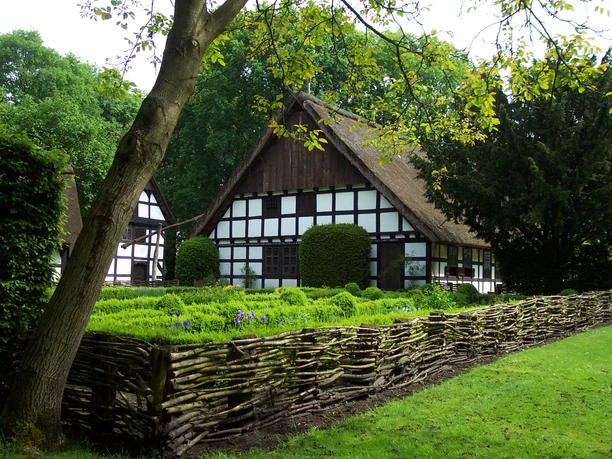BauerngartenMuseumshof.JPG Historisches Fachwerkhaus mit gepflegtem Bauerngarten und Weidenflechtzaun, umgeben von Bäumen.