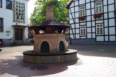 Glindower Platz Rahden Runder Backofen mit roten Ziegeln auf einem gepflasterten Platz, umgeben von Fachwerkhäusern.