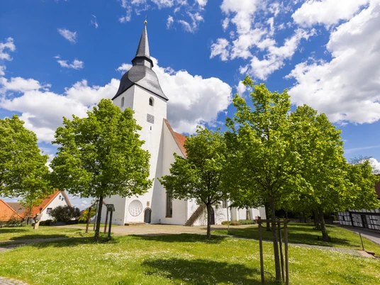 Weiße Stiftskirche Levern mit markantem Turm, umgeben von grünen Bäumen und blauem Himmel.