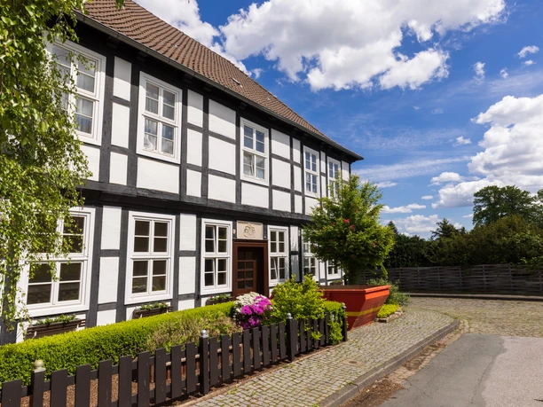 Stiftsort Levern Fachwerkhaus mit Satteldach bei blauem Himmel, umgeben von Blumen und Bäumen an einer gepflasterten Straße.