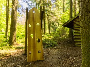 Holzskulptur im Wald mit ausgestanzten Blattformen, neben einer Holzhütte im lichten Forst.
