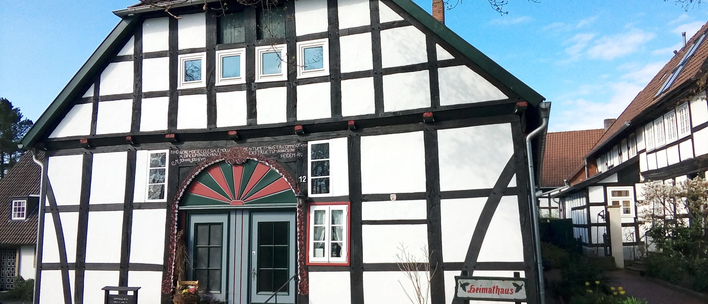 Fachwerkhaus mit markanter Architektur, bunte Tür, gepflegte Vorgartenanlage, blauer Himmel.