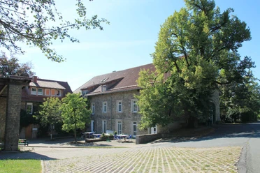 Hof Kaisers Hof Steinbauernhof unter blauem Himmel, von Bäumen flankiert, mit Tischen auf gepflastertem Vorplatz.