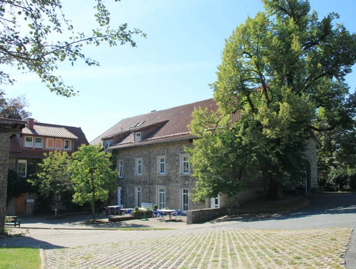 Hof Kaisers Hof Steinbauernhof unter blauem Himmel, von Bäumen flankiert, mit Tischen auf gepflastertem Vorplatz.