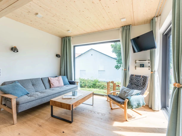 Helles Wohnzimmer mit Holzdecke, grauem Sofa, zwei Sesseln und großem Fenster mit Gartenblick.