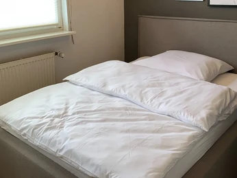 Ein modernes Doppelbett mit weißen Bettbezügen steht in einem schlichten, lichtdurchfluteten Raum.