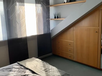 Helles Schlafzimmer unter Dachschräge mit Tageslicht durch Vorhang, Holzschrank und Bett im Hintergrund.
