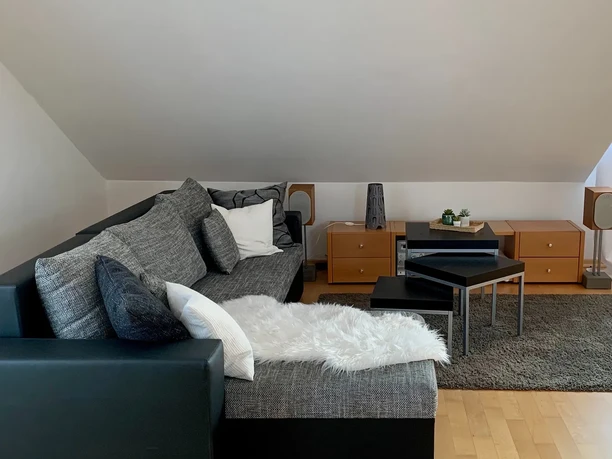 Wohnzimmer Ansicht 2 Modernes Wohnzimmer mit grauem Sofa, Holzboden, Couchtisch, Teppich und dezenter Dekoration.