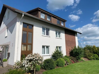 Mehrstöckiges Wohnhaus mit weißer Fassade, braunen Elementen und gepflegtem Garten an sonnigem Tag.