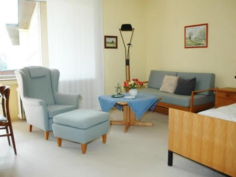 Helles Zimmer mit Sessel, Ottomane, Sofa, Tisch und Bett in sanften Farbtönen, Fenster links.