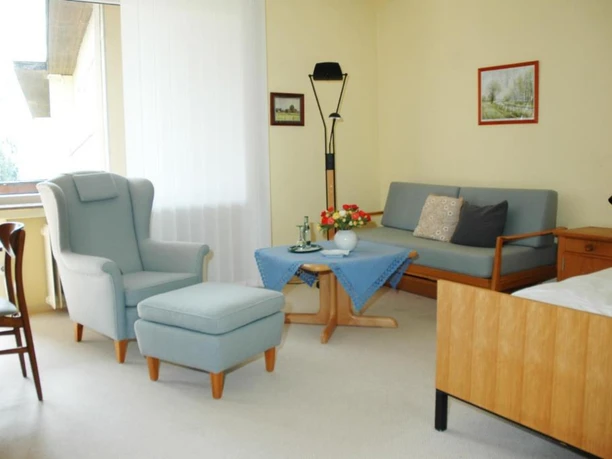 Zimmer 2 Helles Zimmer mit Sessel, Ottomane, Sofa, Tisch und Bett in sanften Farbtönen, Fenster links.