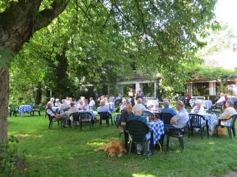 Gäste sitzen entspannt auf Plastikstühlen an blau-weiß karierten Tischen unter schattigen Bäumen.