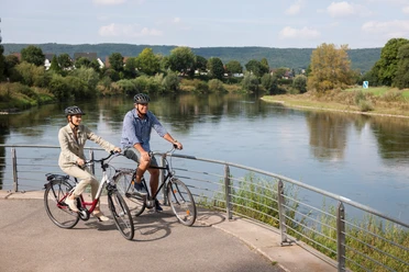 Zwei Radfahrer stehen auf einer Brücke, die über einen Fluss mit bewaldeten Ufern führt.