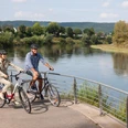 Werre-Weser-Kuss Zwei Radfahrer stehen auf einer Brücke, die über einen Fluss mit bewaldeten Ufern führt.