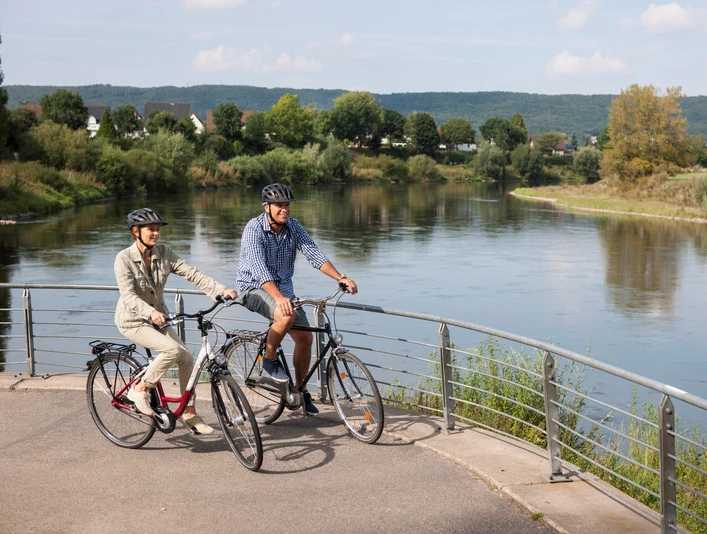 Werre-Weser-Kuss Zwei Radfahrer stehen auf einer Brücke, die über einen Fluss mit bewaldeten Ufern führt.