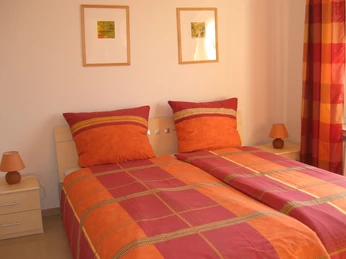 Schlafzimmer Zwei Einzelbetten mit orange-roter Bettwäsche, helle Möbel, zwei Wandbilder, je eine Lampe pro Nachttisch.