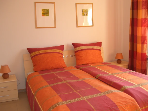 Schlafzimmer Zwei Einzelbetten mit orange-roter Bettwäsche, helle Möbel, zwei Wandbilder, je eine Lampe pro Nachttisch.