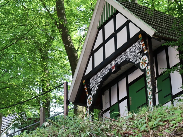 Kahle Wart Fachwerkhäuschen im Wald, umgeben von grünem Laub, auf einem Hügel gelegen, malerische Szene.