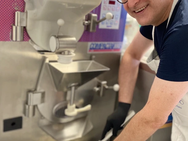 Mann in Eisdiele bereitet frisch Gelato zu, lächelt in die Kamera, trägt Schürze und schwarze Handschuhe.