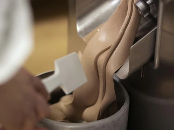 Eine Person füllt frisch zubereitetes Schokoladeneis aus einer Eismaschine in einen Metallbehälter.