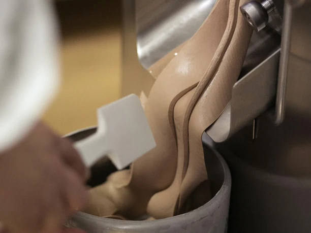 Eine Person füllt frisch zubereitetes Schokoladeneis aus einer Eismaschine in einen Metallbehälter.