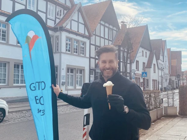 Ein Mann steht auf einer gepflasterten Straße, hält ein Eis und lächelt in die Kamera, neben ihm ein blaues Werbebanner. Im Hintergrund sind malerische Fachwerkhäuser und ein klarer Himmel zu sehen.