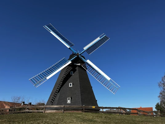 Eine schwarze Windmühle mit weißen Flügeln erhebt sich majestätisch vor strahlend blauem Himmel.