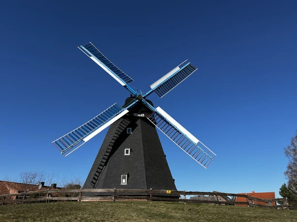 Eine schwarze Windmühle mit weißen Flügeln erhebt sich majestätisch vor strahlend blauem Himmel.