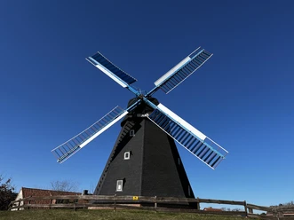 Windmühle Paula mit schwarzen Flügeln und tiefblauem Himmel im Hintergrund, umgeben von Holzzaun.