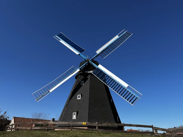 Windmühle Paula Windmühle Paula mit schwarzen Flügeln und tiefblauem Himmel im Hintergrund, umgeben von Holzzaun.
