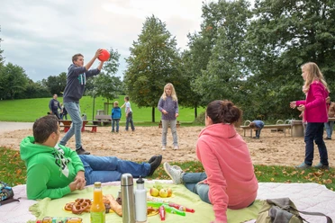 Freizeitpark Langfort in Langenfeld Menschen entspannen auf einer grünen Wiese, während Kinder im Hintergrund Ballspiele auf einem Spielplatz genießen.