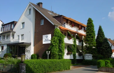 Ein dreistöckiges Gebäude mit weißer Fassade und braunen Balkonen, umgeben von Pflanzen und Bäumen.