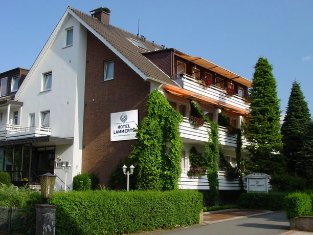 Ein dreistöckiges Gebäude mit weißer Fassade und braunen Balkonen, umgeben von Pflanzen und Bäumen.