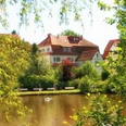 Haus mit rotem Dach am See, umgeben von Bäumen und Spazierweg; Schwan auf dem Wasser.
