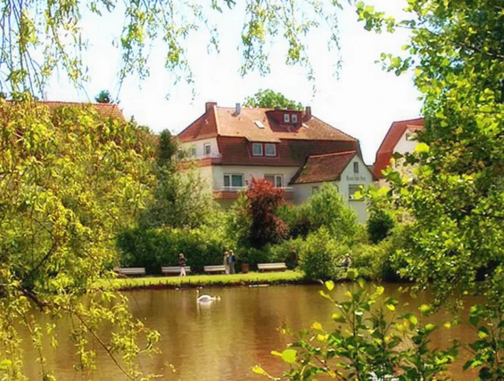 Haus am See Haus mit rotem Dach am See, umgeben von Bäumen und Spazierweg; Schwan auf dem Wasser.