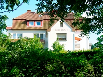 Großes weißes Gebäude mit rotem Dach, umgeben von dichten grünen Bäumen, bei sonnigem Wetter.