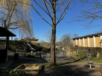 Wasser+Mehr Erlebnispark Teich mit Steg im Vordergrund, von Bäumen umgeben, blauer Himmel und Gebäude im Hintergrund.
