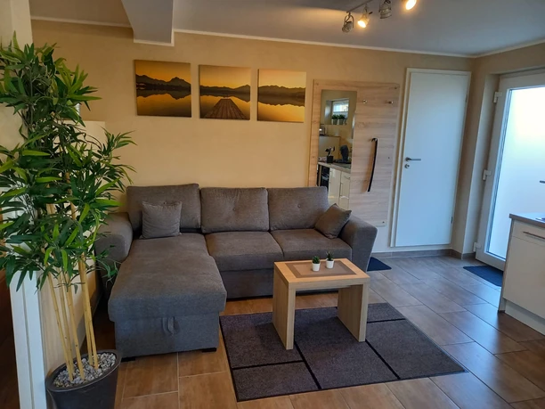 City-Apartment Zum Alten Graf_Wohnraum Gemütliches Wohnzimmer mit grauem Ecksofa, Holzcouchtisch, Pflanzen, Wandbildern und hellen Fliesen.