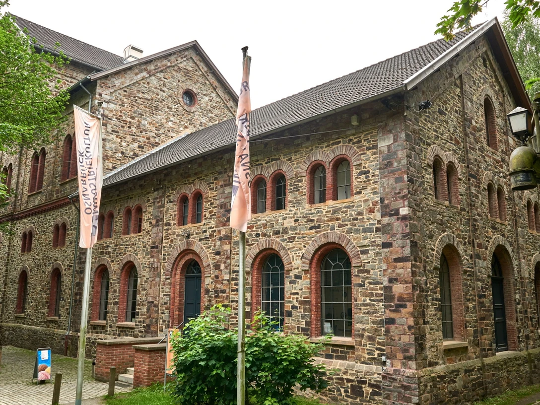 Das MIK von Außen Historisches Backsteingebäude mit spitzem Dach und großen Fenstern, umgeben von Bäumen und Weg.Historic brick building with a pointed roof and large windows, surrounded by trees and a path.Historisch bakstenen gebouw met een puntdak en grote ramen, omringd door bomen en een pad.