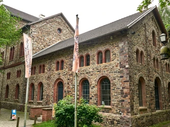 Das MIK von Außen Historisches Backsteingebäude mit spitzem Dach und großen Fenstern, umgeben von Bäumen und Weg.Historic brick building with a pointed roof and large windows, surrounded by trees and a path.Historisch bakstenen gebouw met een puntdak en grote ramen, omringd door bomen en een pad.Historisk murstensbygning med spidst tag og store vinduer, omgivet af træer og en sti.