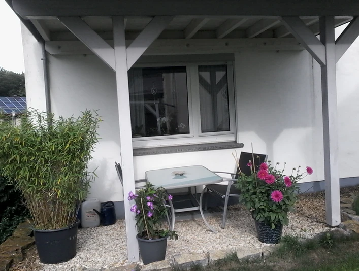 Ferienwohnung Carpe Diem Außenbereich Überdachte Terrasse der Ferienwohnung Carpe Diem mit Tisch, Stühlen und Topfpflanzen in blühender Umgebung.